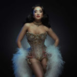 Jolie Papillon with her hands on her waist and wearing a golden corset and white feather boa