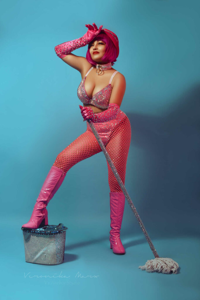 Burlesque star Cleopantha in pink, holding a mop and with one foot on a metal bucket.