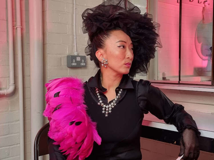 Fancy Chance seated in a dressing room wearing a black headdress and with a hot pink shoulder detail.