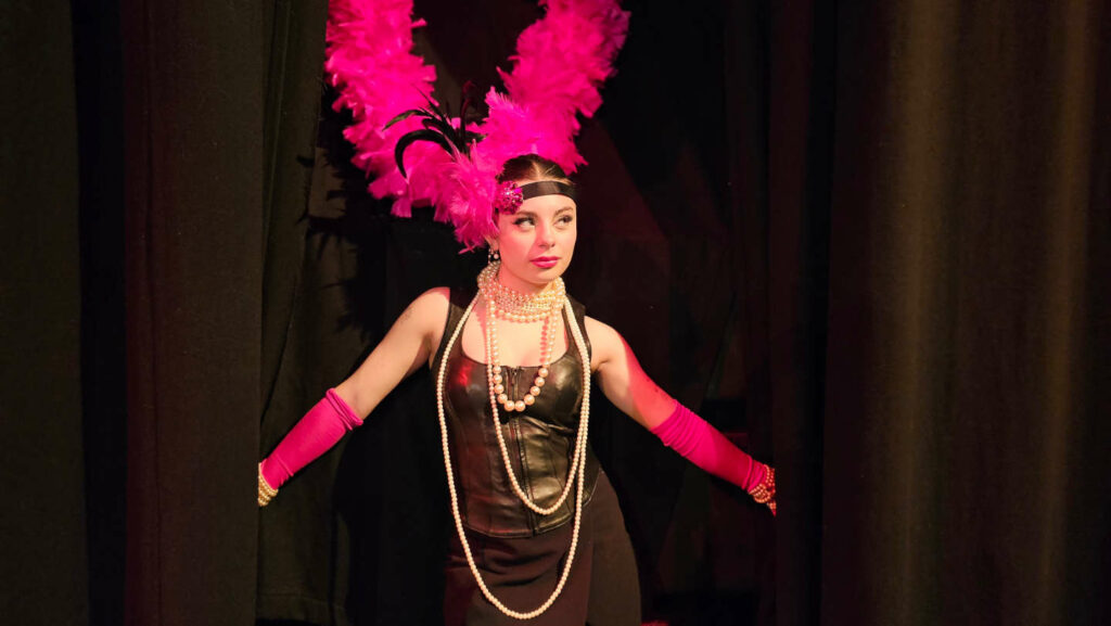 Sabrina King wearing hot pink feathers and evening gloves poses in front of a black curtain in a theatre.