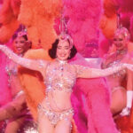 Dita Von Teese in a silver showgirl costume surrounded by showgirl performers with pink and orange feathers